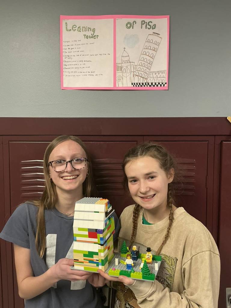 Two middle school students smiling and holding their LEGO project, a tall white and yellow tower. Behind them on a maroon locker is a poster titled 'Leaning Tower of Pisa' that includes a detailed drawing and facts about the landmark.