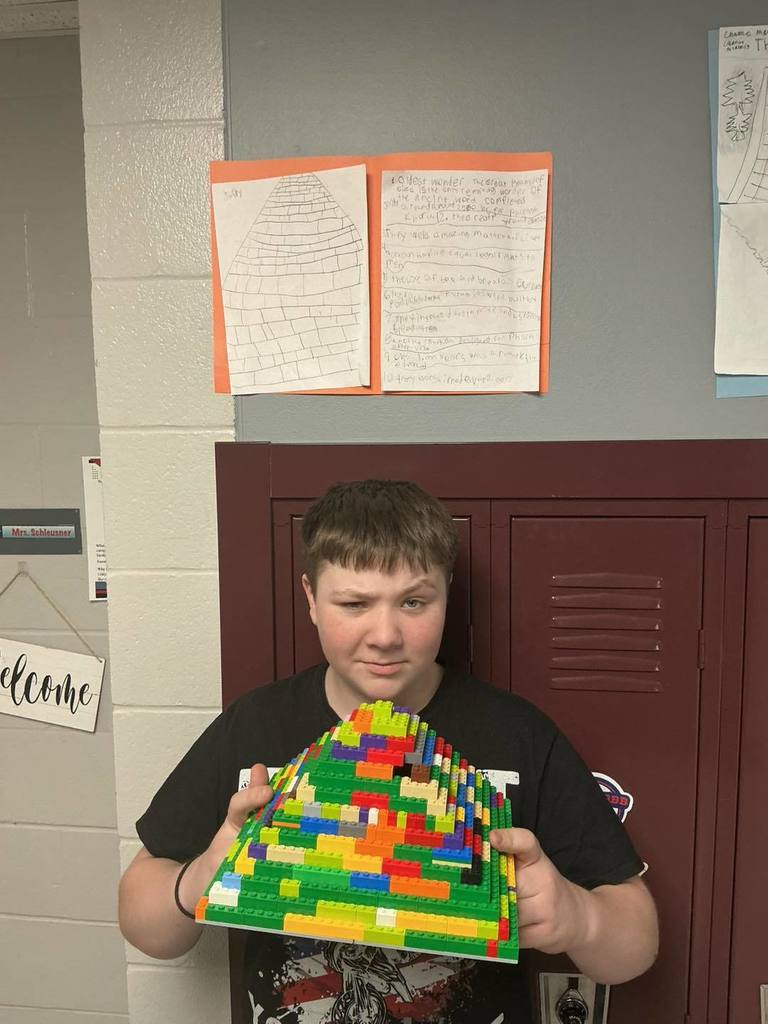 A middle school student holding a large, multi-colored LEGO pyramid project in front of his locker. Above him, a poster titled 'Great Pyramid of Giza' is displayed, featuring a large drawing of a pyramid and handwritten research notes.