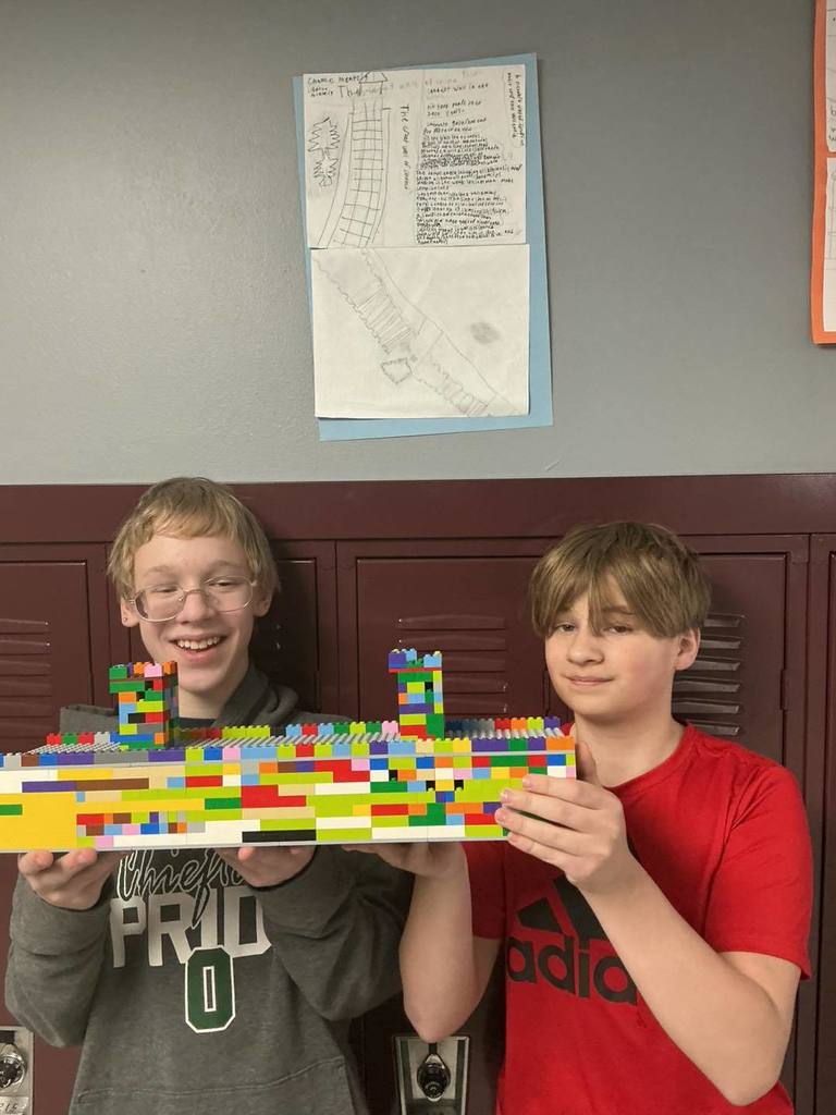 Two middle school students smiling while holding a colorful LEGO house they built for their landmark project. Behind them, a poster titled 'The Great Wall of China' is displayed on a locker, featuring facts and illustrations of the landmark.
