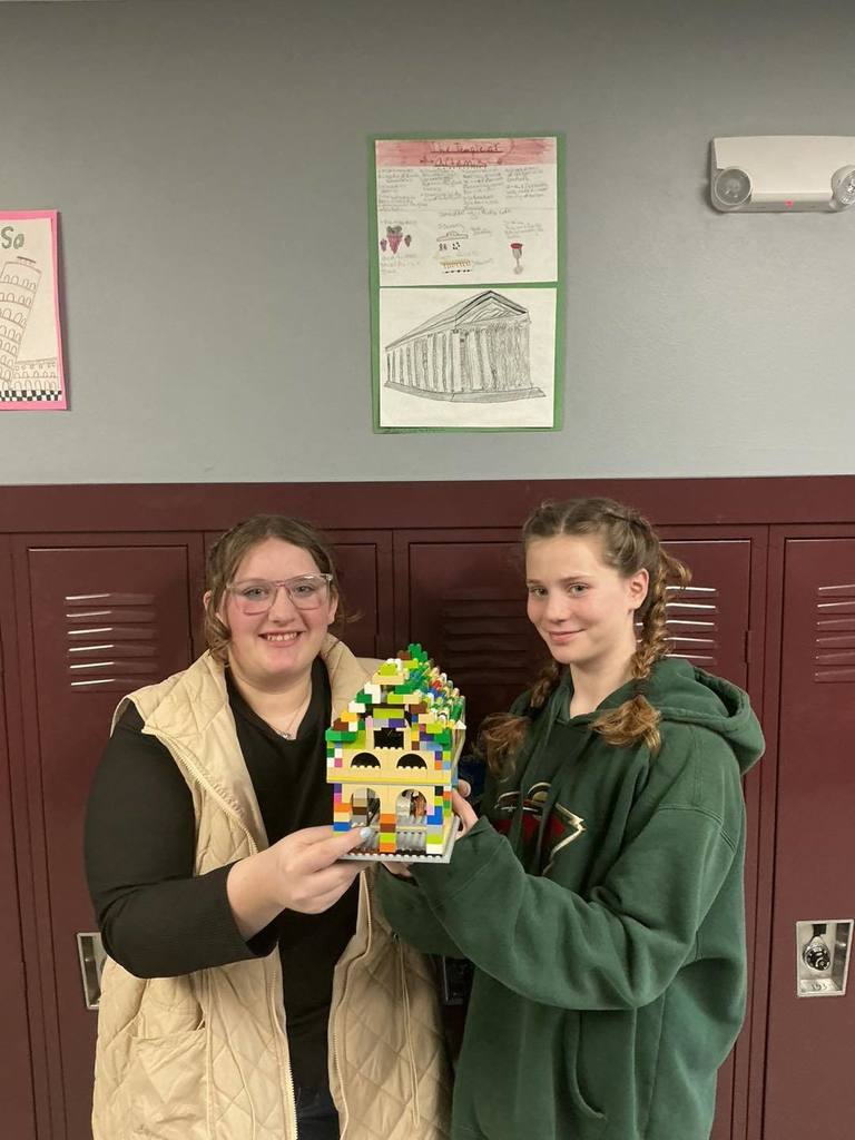 Two middle school students smiling and holding a green and yellow LEGO building. Behind them on the locker is a poster titled 'The Parthenon' featuring a large drawing of the ancient Greek temple and historical facts.