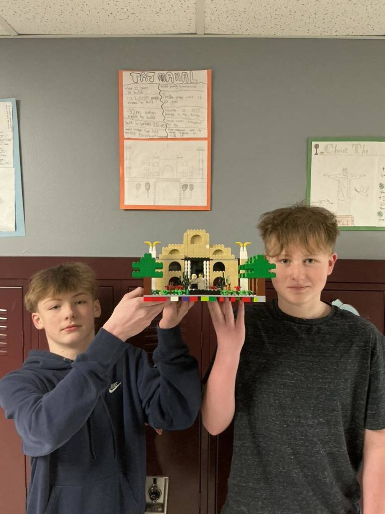 Two students posing with their LEGO model of the Taj Mahal. Behind them on the classroom wall, a detailed poster titled 'The Taj Mahal' is displayed, featuring facts and diagrams about the iconic landmark.