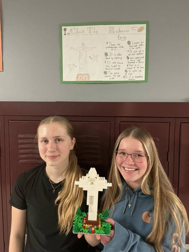 Two middle school students from Unity School District posing with their LEGO model of Christ the Redeemer. Behind them, a poster titled 'Christ the Redeemer' is displayed on a locker, featuring a drawing of the statue and bulleted facts about the landmark.