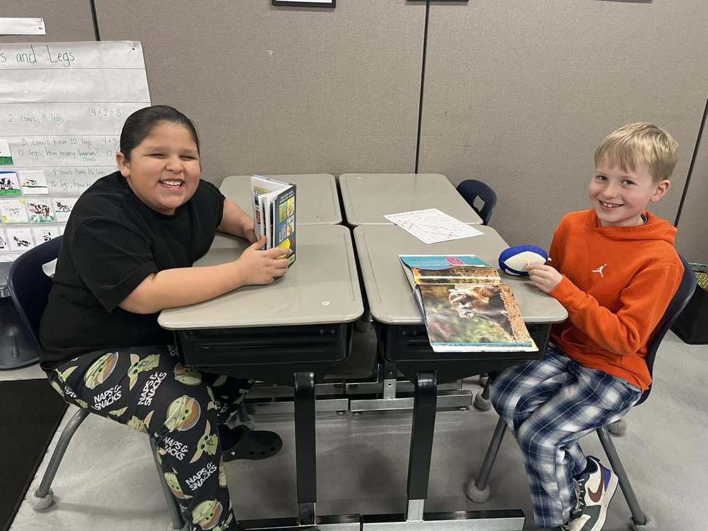 Two third-grade students at Unity School District smiling at their desks during Read Across America Week. One student is wearing 'Star Wars' themed pajamas and holding a book, while the other is in an orange sweatshirt with blue plaid pajama pants, with several books open in front of them.