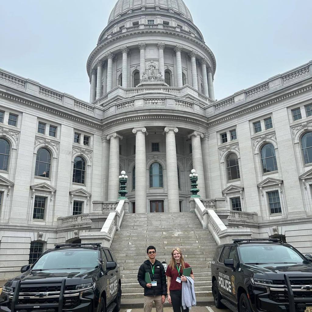 Two Unity High School students standing together on the steps of the Wisconsin State Capitol in Madison. The massive white granite dome and ornate architectural details of the Capitol building rise high into the sky behind them.