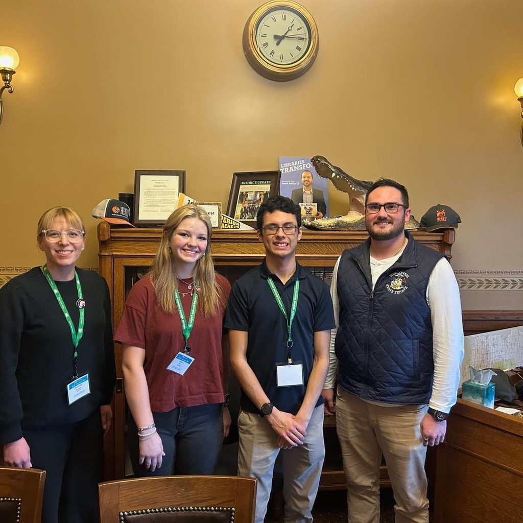 Two Unity High School students and an adult advisor posing for a photo in a legislator's office at the Wisconsin State Capitol. They are standing in front of a large wooden desk with a clock and various official documents on the wall behind them.