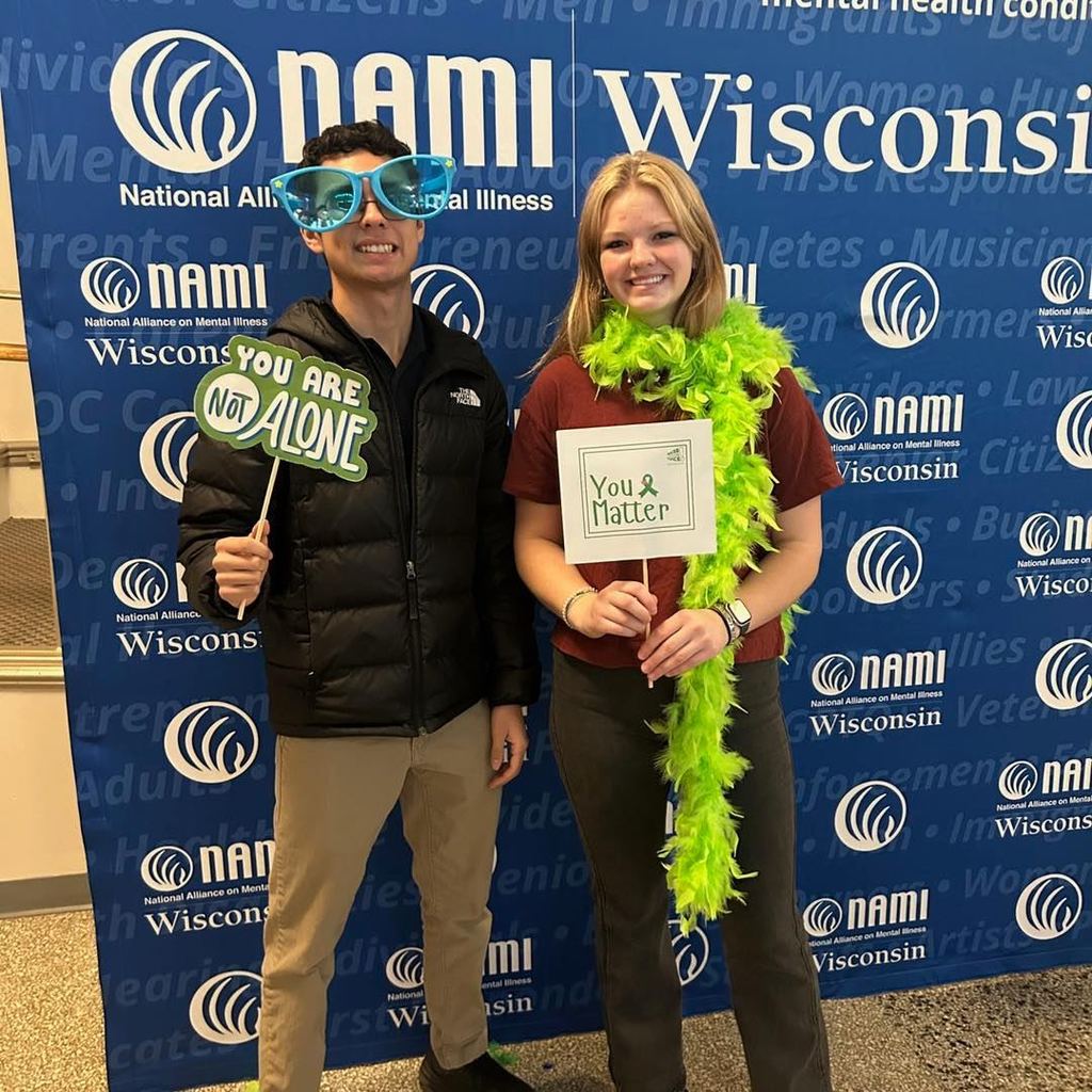 Two Unity High School students posing in front of a NAMI Wisconsin backdrop. One student is wearing a green feather boa and holding a sign that says 'You Matter,' while the other holds a sign that says 'You Are Not Alone.