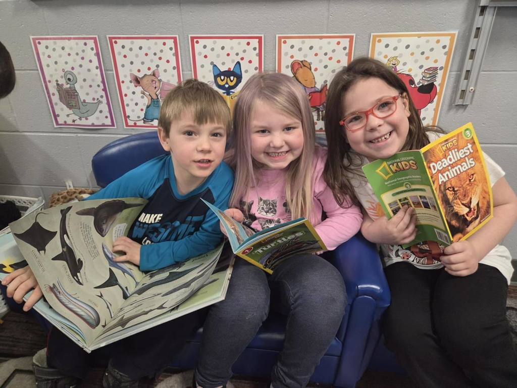 Three Unity elementary students sitting together on a blue beanbag chair during Read Across America Week. They are smiling and showing off their books about sharks, turtles, and deadly animals.