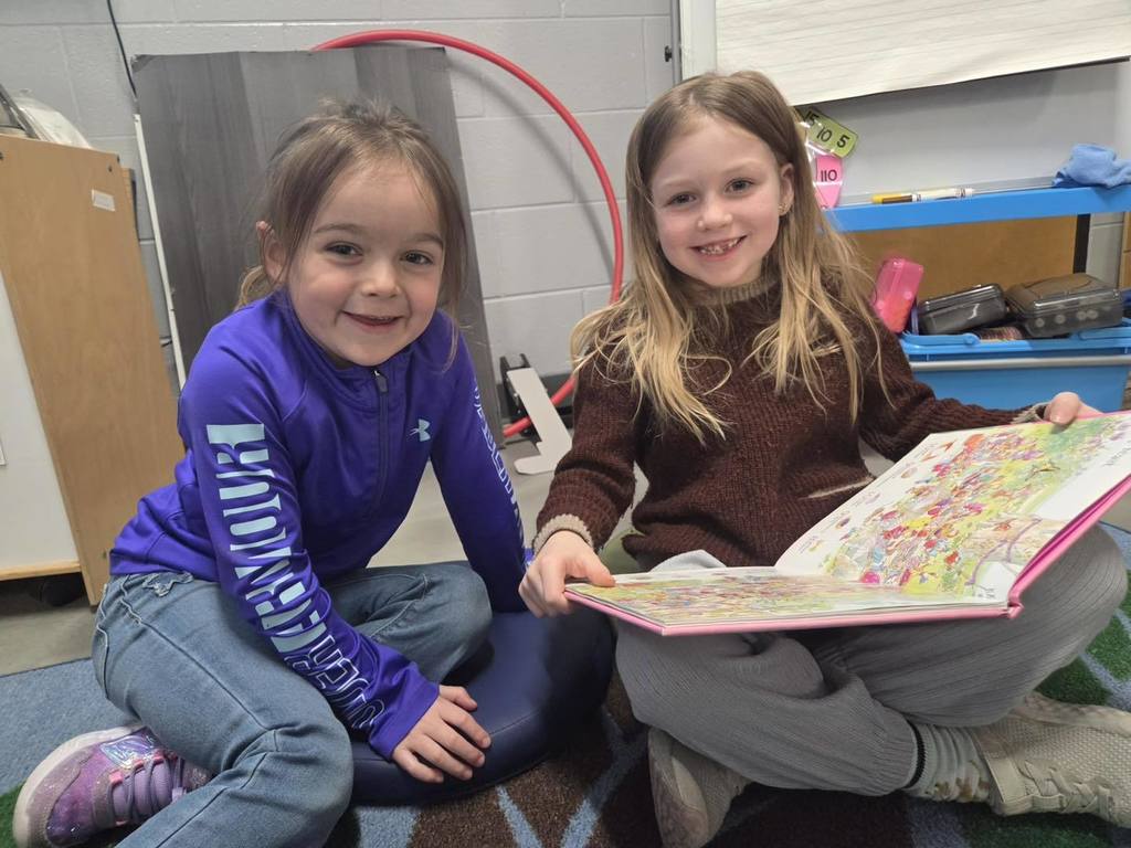Two elementary students sitting on a classroom rug, smiling while sharing a large, colorful picture book during Read Across America Week. One student is wearing a purple Under Armour sweatshirt, and the other is in a brown sweater.
