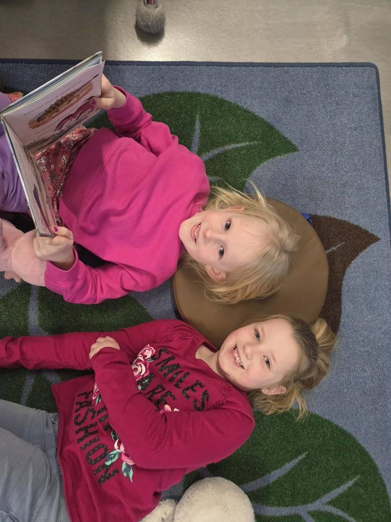 Two elementary students in pink outfits laying on a leaf-patterned rug during Read Across America Week. They are smiling at the camera while sharing a book together in a cozy classroom reading nook.