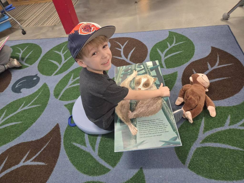 A Unity elementary student wearing a blue and orange baseball cap, sitting on a leaf-patterned rug. He is smiling while holding an open book featuring a large photo of a slow loris, with a small stuffed monkey sitting on the rug next to him.