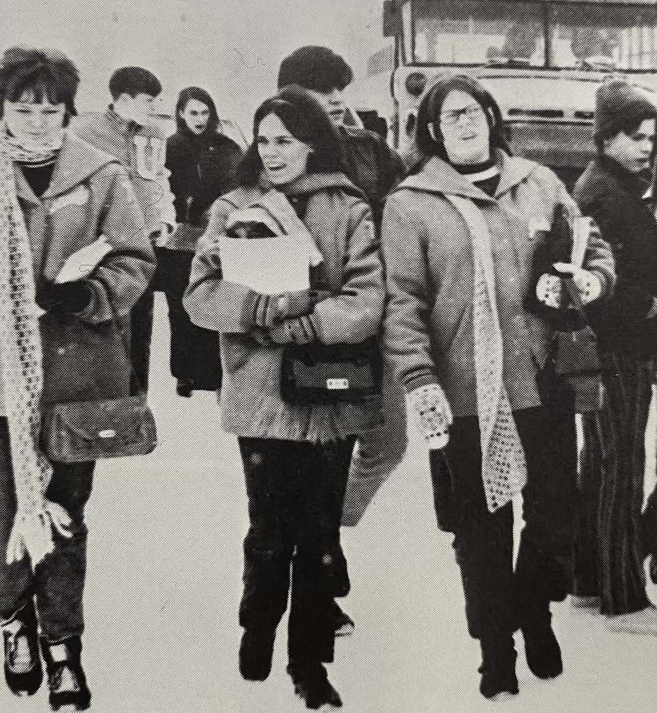A vintage black-and-white photo of four Unity students walking outdoors together in winter clothing. They are carrying books and binders, with a school bus visible in the background, capturing a candid moment from a past school year.