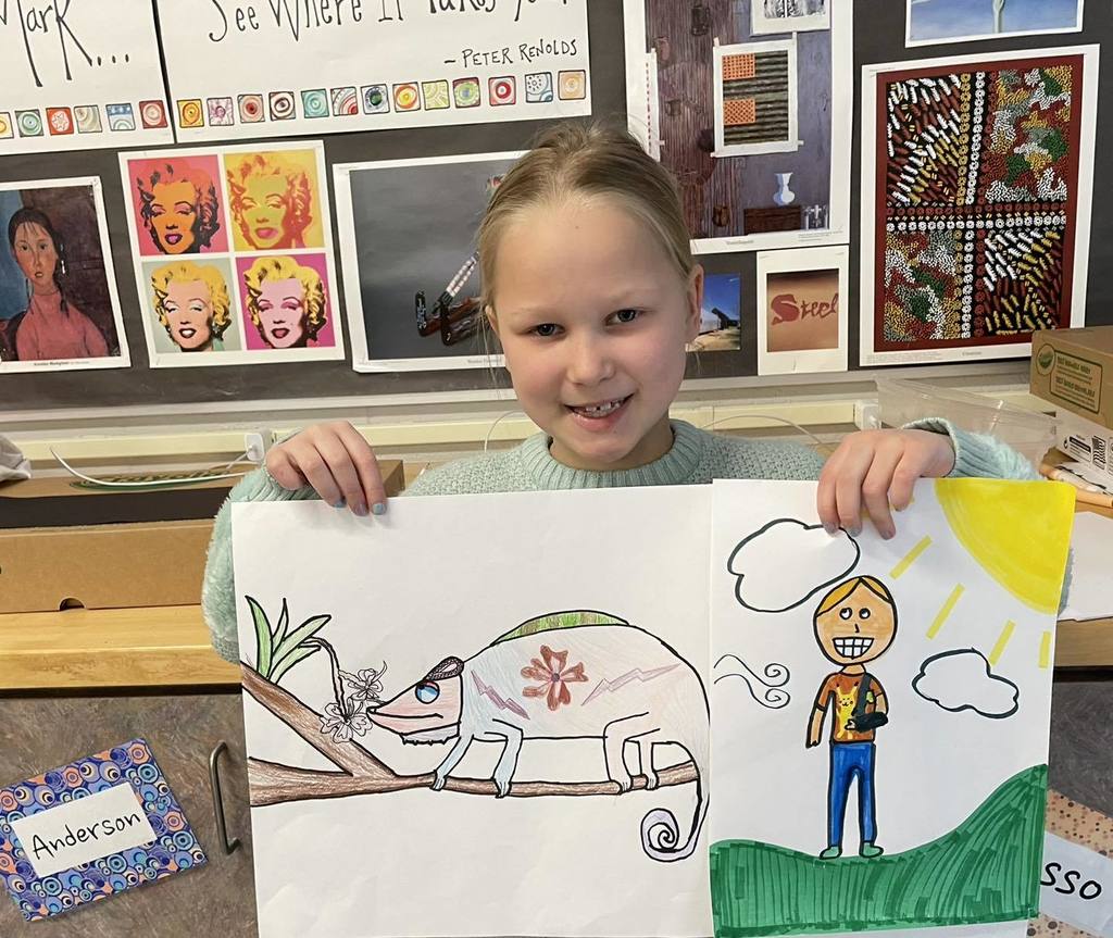 A Unity third-grade student named Fiona smiling and holding up two colorful drawings. One drawing features a detailed, patterned chameleon on a branch, and the other shows a person standing under a bright yellow sun. She is standing in front of a classroom wall decorated with various student art pieces.