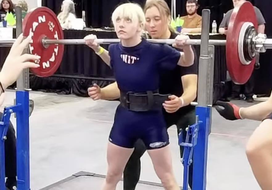 A Unity powerlifter with blonde hair performing a squat at the State meet. She is wearing a blue 'Unity' singlet and a lifting belt, with a coach spotting her from behind and heavy weights loaded on the barbell.