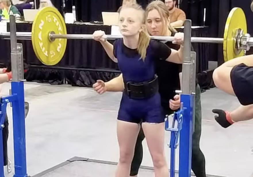 A Unity powerlifter with blonde hair tied back, wearing a blue Unity singlet and lifting belt, performing a squat at the State competition. A female coach stands directly behind her, ready to spot the lift, in a crowded gym environment with yellow 'Titan' weight plates on the barbell.