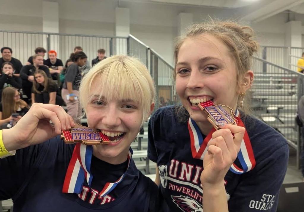 A close-up of Unity powerlifters Khloe Jensen and Persayiss Tucker smiling and celebrating their 5th-place State medals. They are both wearing their medals and biting them playfully for the camera, dressed in their Unity Powerlifting gear with the competition bleachers in the background.