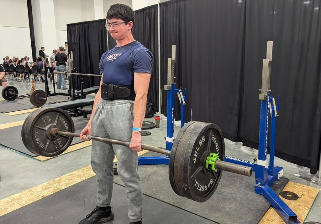 Unity powerlifter Marius Bruce standing over a loaded barbell, preparing for a deadlift at the State meet. He is wearing a blue Unity t-shirt, grey sweatpants, and a lifting belt, with a focused expression as he prepares to pull the weight.
