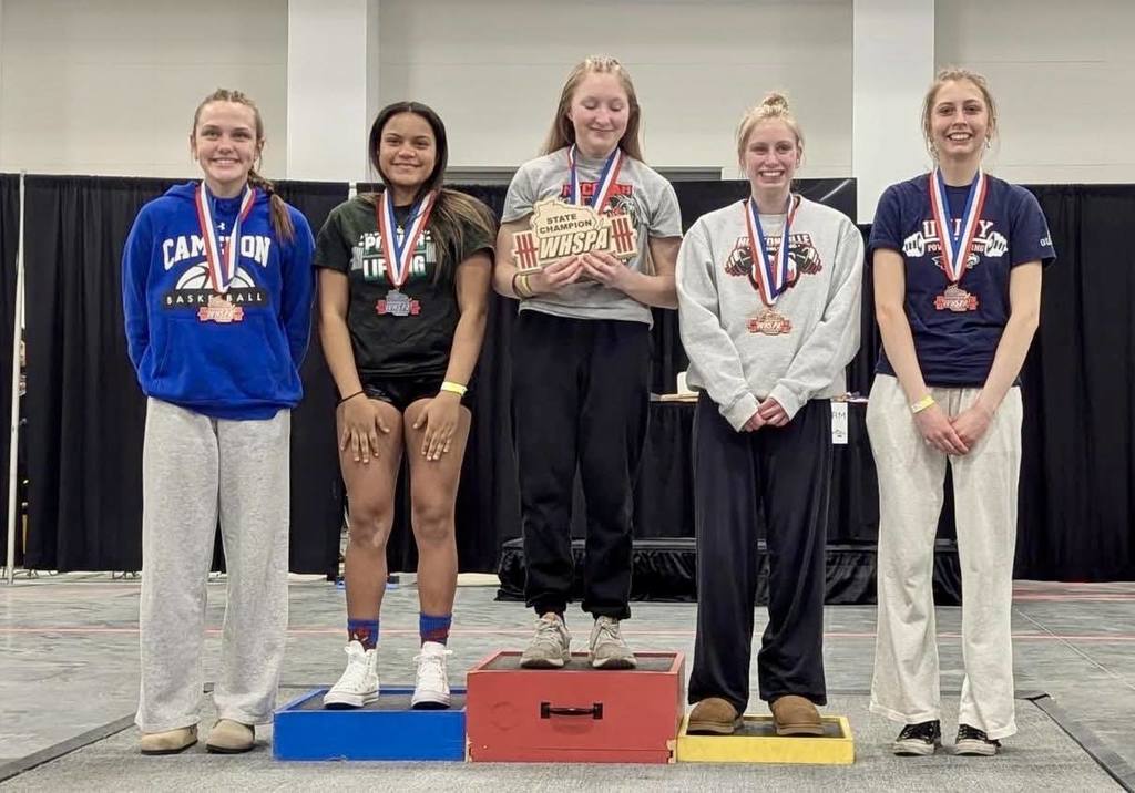 For the third photo in your set (the podium shot), here is the alt text: "Five female powerlifters standing on a tiered victory podium at the State meet. A Unity student in a grey 'State Champion' shirt stands on the highest central platform, while four other medalists, including a Unity student in a blue sweatshirt, stand on the surrounding steps wearing their medals.
