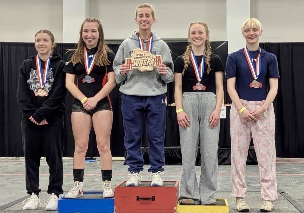 Five female powerlifters standing on a tiered victory podium at the State meet. A Unity student stands on the far right in a pink-patterned pair of pants, wearing her 5th-place medal. In the center, a lifter in a grey sweatshirt holds a 'State Champion' plaque.