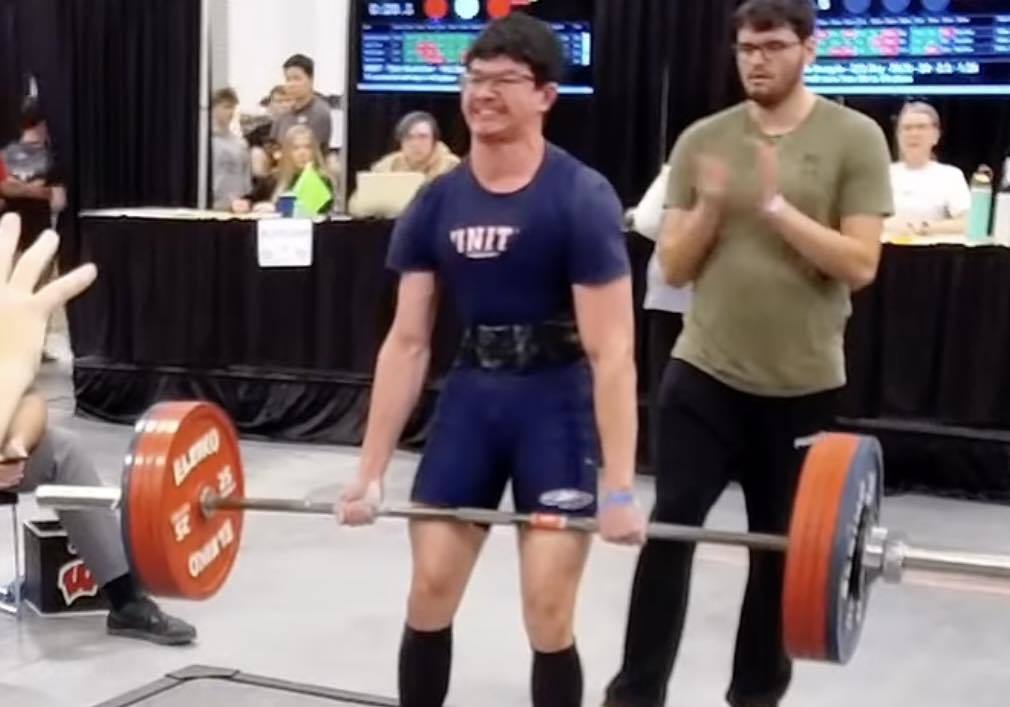 A Unity powerlifter, Marius Bruce, performing a deadlift at the State meet. He is wearing a blue Unity t-shirt and a lifting belt, gripping a heavily loaded barbell. A coach stands behind him, clapping and cheering him on as he completes the lift.