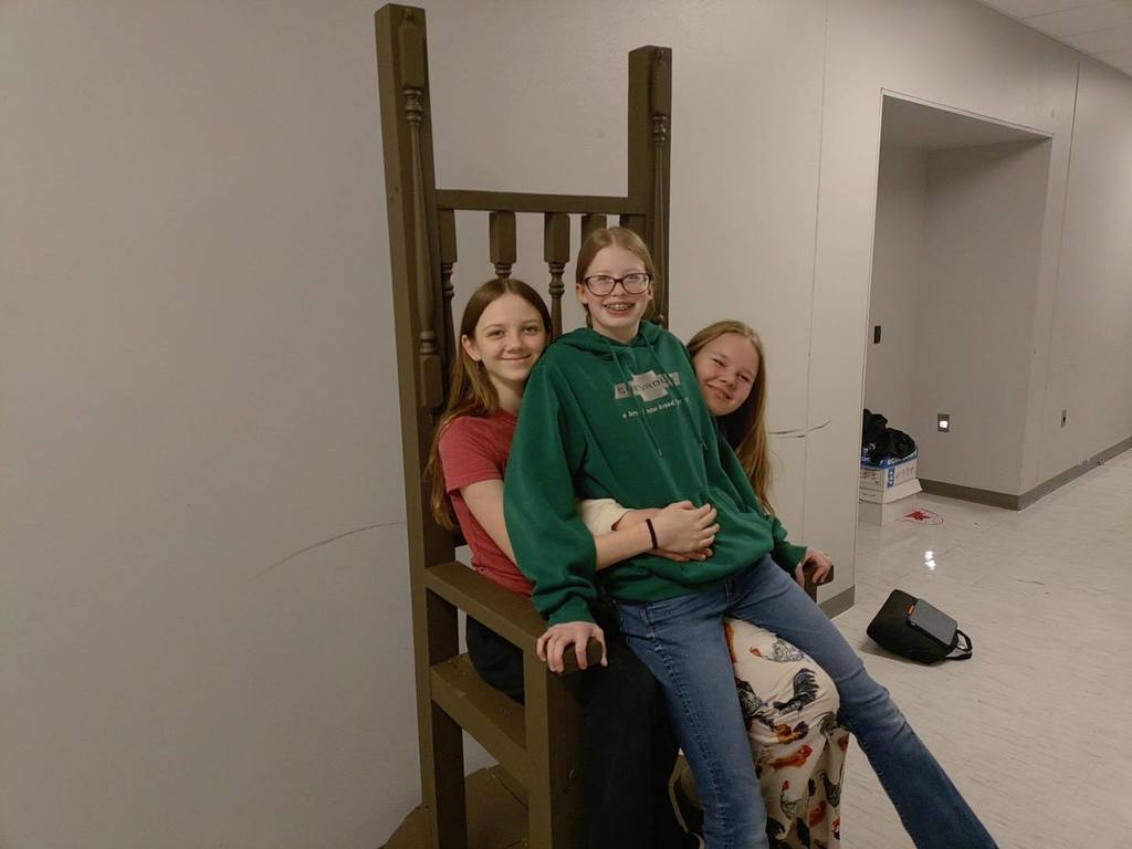 Three Unity students posing together on and around a large, ornate wooden throne built for a stage production. One student sits in the tall chair while two others lean against it, smiling in a school hallway to showcase the completed set piece.