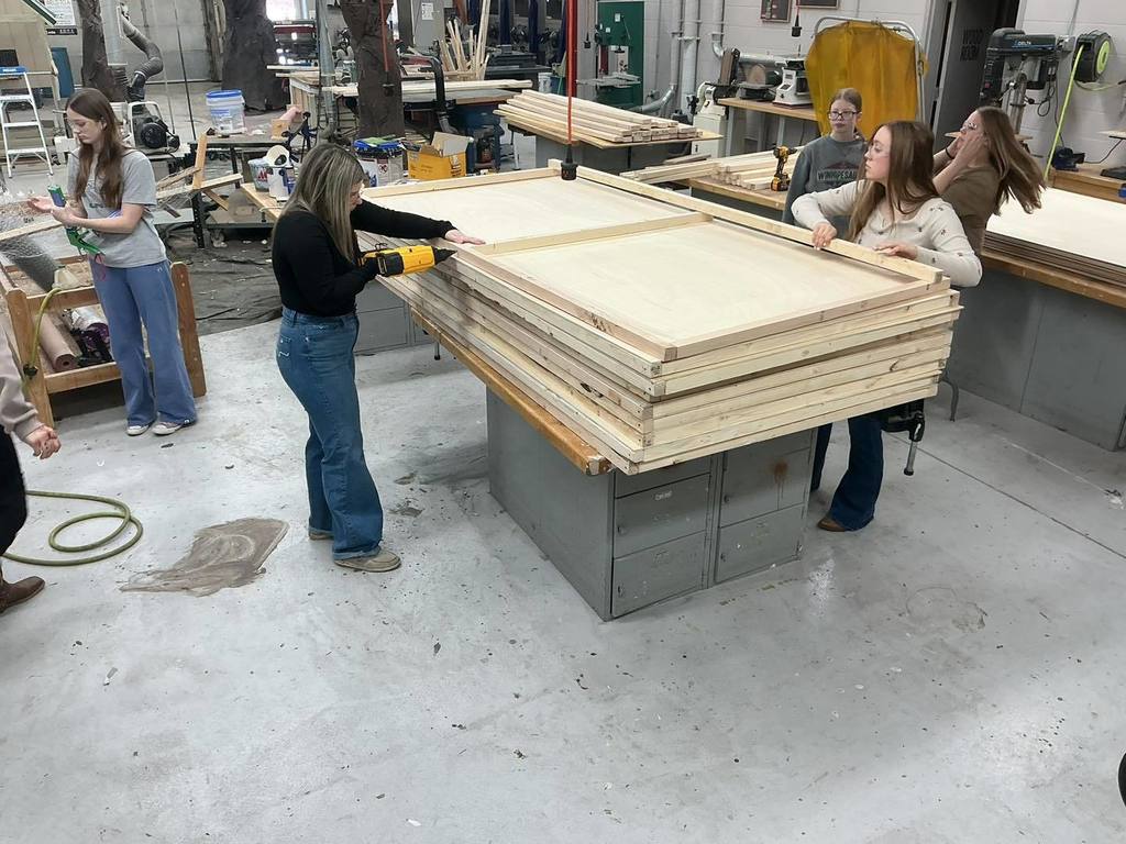 Four Unity students working together in a woodshop to build a large stage platform. One student uses a power drill to secure a wooden frame, while the others hold the large plywood structure in place on a workbench, surrounded by various tools and machinery.