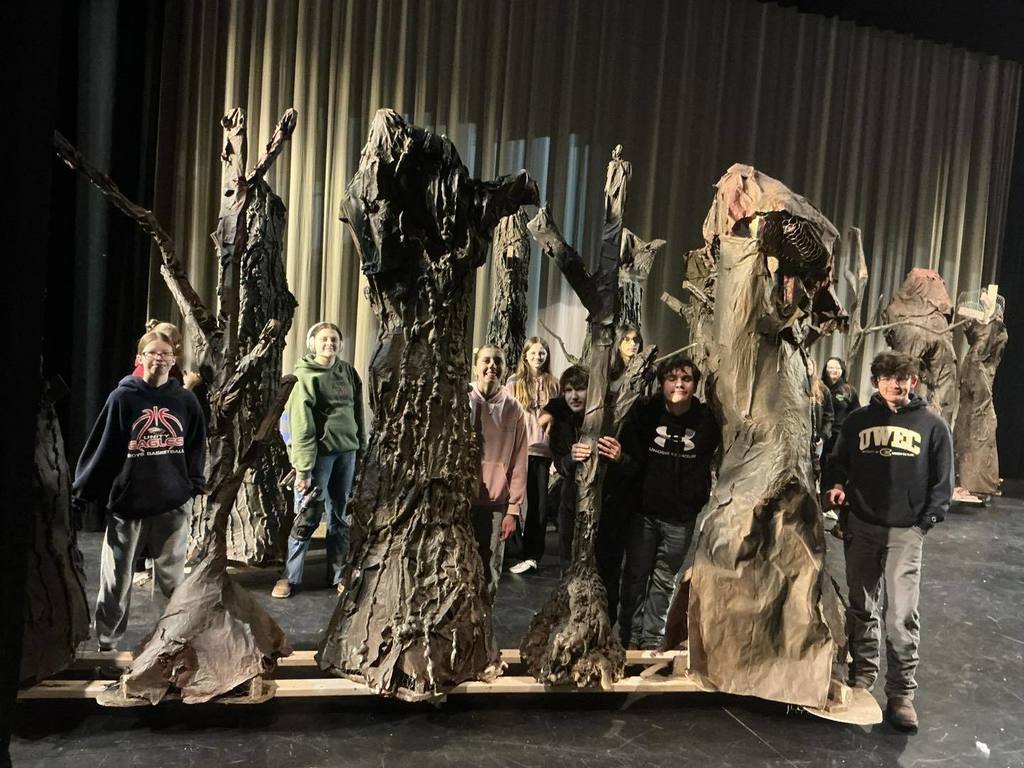 A group of Unity Set Design students standing on a stage behind several tall, hand-crafted tree trunk set pieces. The students are smiling among their work, which features realistic textures and branches, created for the upcoming productions of Frozen, Robinhood, and Annie Junior.