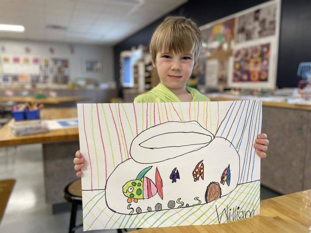 A young student named William smiling and holding up his artwork in a classroom. His drawing features a large, colorful creature with patterns, surrounded by smaller figures and vertical rainbow-colored stripes. His name, 'William,' is written in the bottom right corner of the paper.