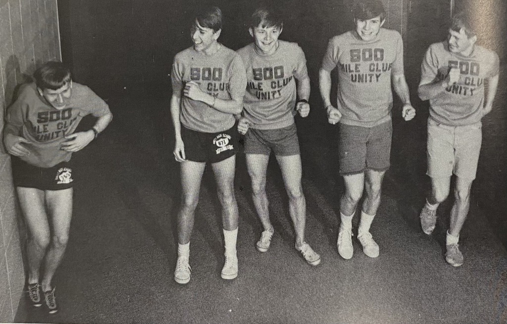 A black and white throwback photo of five Unity students in athletic gear. They are mid-stride, pretending to run toward the camera. Each student is wearing a t-shirt that says '500 Mile Club Unity' with a winged foot logo. One student on the far left is leaning against a wall in a running pose.