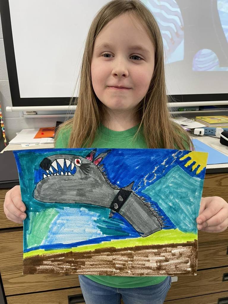 A young student smiling and holding up her colorful drawing of a gray shark with sharp teeth swimming in a blue ocean under a bright yellow sun.