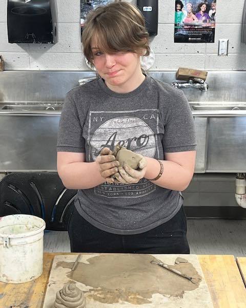 A high school student in a gray "Aero" t-shirt stands at a classroom workbench, smiling while holding a piece of gray clay. On the table in front of her is a flattened slab of clay and a sculpting tool, with a white bucket nearby.