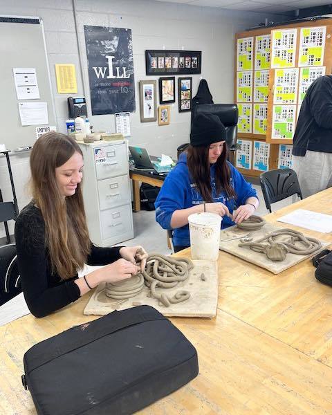 Two high school students sit at a classroom table working with clay. They are using their hands to shape long, thick clay coils into circular patterns on wooden boards.