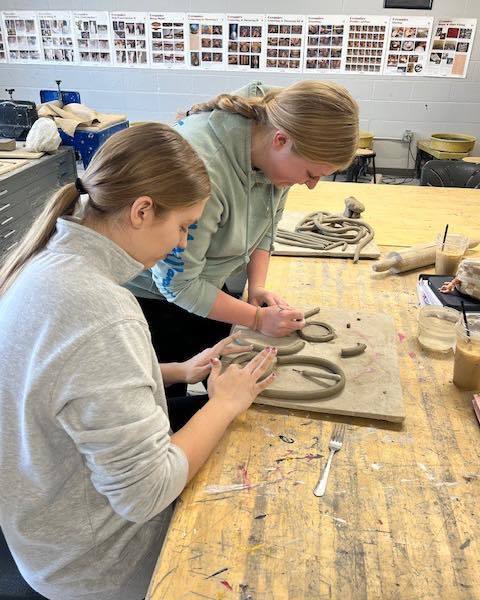 Two high school students working at a wooden table in an art classroom, carefully shaping long coils of gray clay into circular designs on wooden boards.
