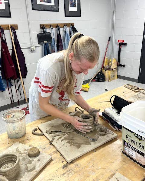 A high school student with a blonde ponytail smiles as she works at an art station, using her hands to carefully stack and smooth clay coils to build the walls of a ceramic vessel.