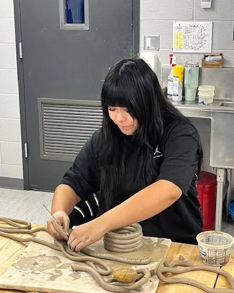 A high school student with long dark hair wearing a black shirt works on a ceramics project. She is carefully layering thick clay coils to build a vessel, with several more coils prepared on the table in front of her.