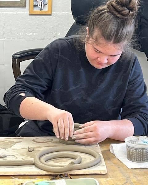 A high school student with her hair in a bun leans over a wooden table, using her hands to carefully press and join a long clay coil onto the base of a ceramic project.