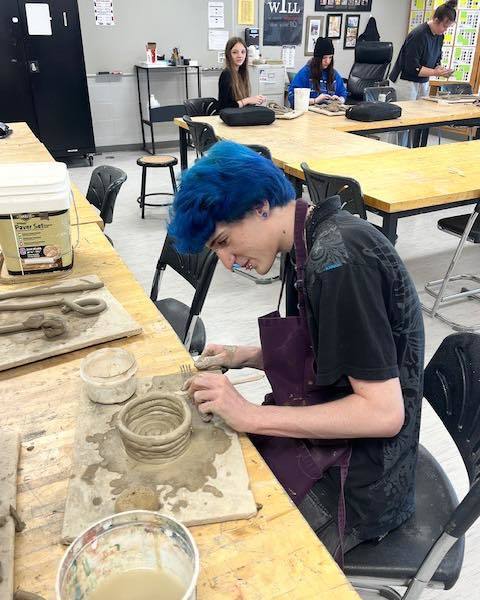 A high school student with bright blue hair wearing a maroon apron, using a metal tool to carefully score and join clay coils to build a ceramic pot in an art classroom.