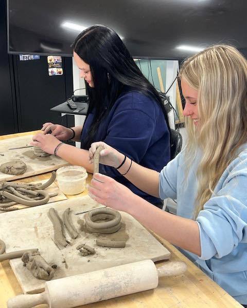 Two high school students working side-by-side at a classroom table. One student in a blue sweatshirt is carefully lifting a long clay coil, while the student next to her in a dark shirt focuses on shaping her own ceramic piece.