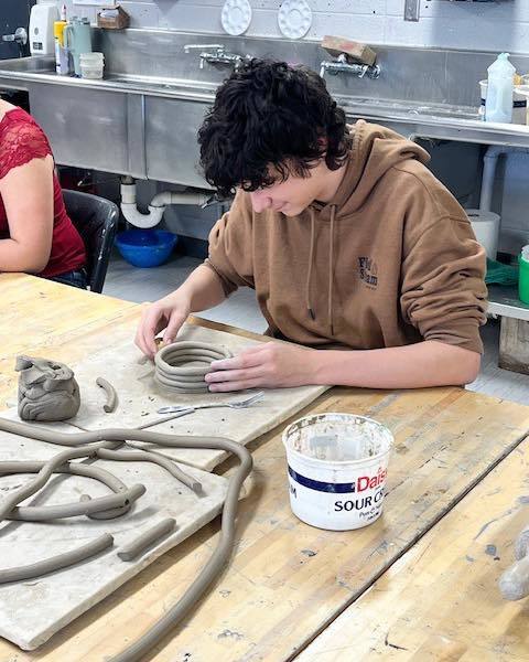 A high school student in a brown hoodie sits at a wooden workbench, carefully stacking gray clay coils to build a vessel. Several long, hand-rolled clay coils and a small white container sit on the table in front of him.