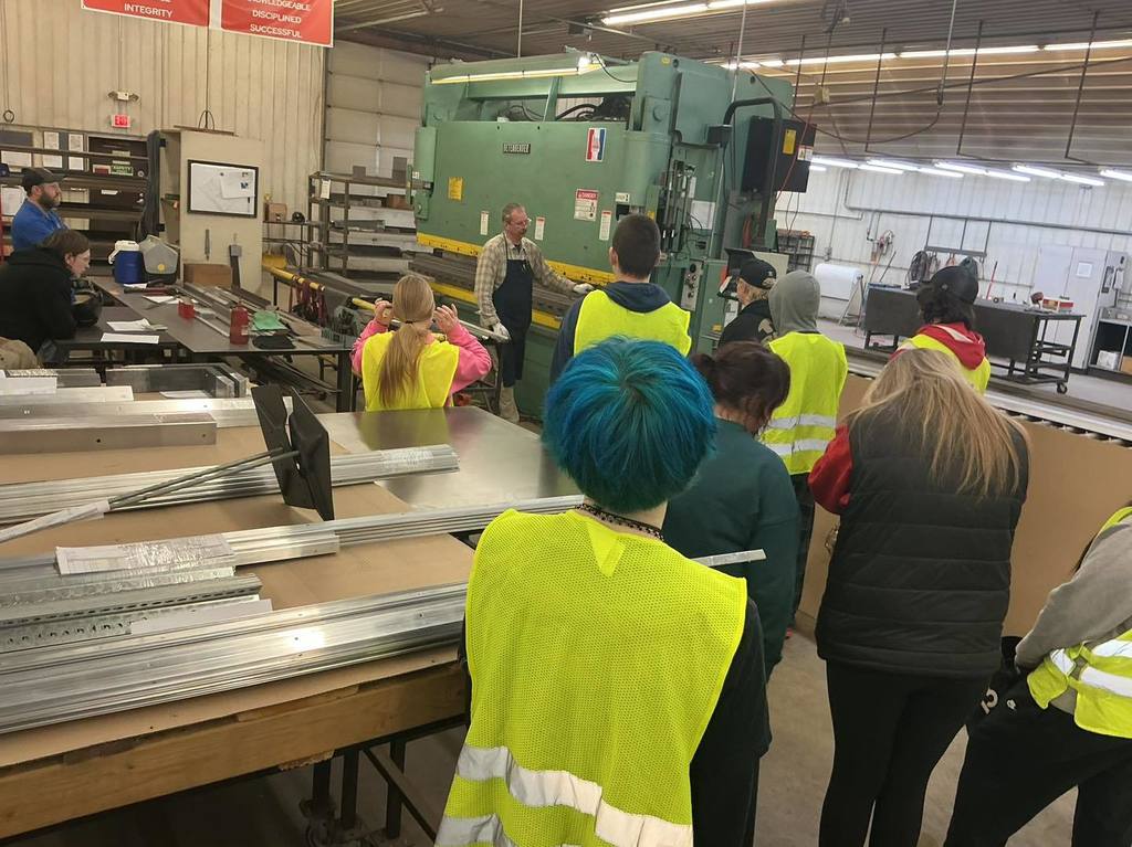 A group of high school students in yellow safety vests stand in a circle inside a large manufacturing shop. They are watching a demonstration of a large green industrial press machine led by a staff member at Bishop Fixtures.