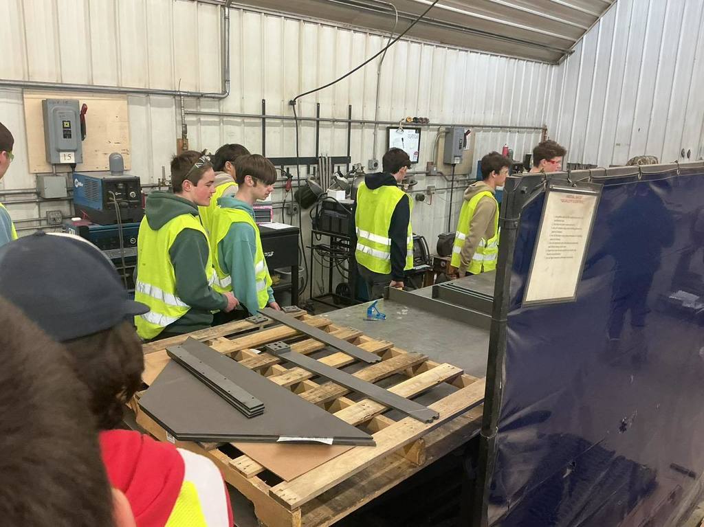 A group of high school students in yellow safety vests stand around a wooden pallet in a manufacturing shop. They are observing several flat, dark gray metal components that were fabricated for their custom toolbox kits.