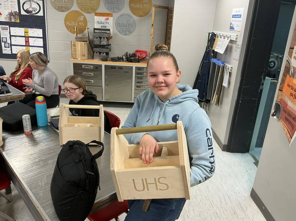 A high school student in a light blue hoodie sits at a classroom desk, smiling and holding a custom wooden toolbox with "UHS" engraved on the side. Another student and several more wooden toolboxes are visible on the tables in the background.