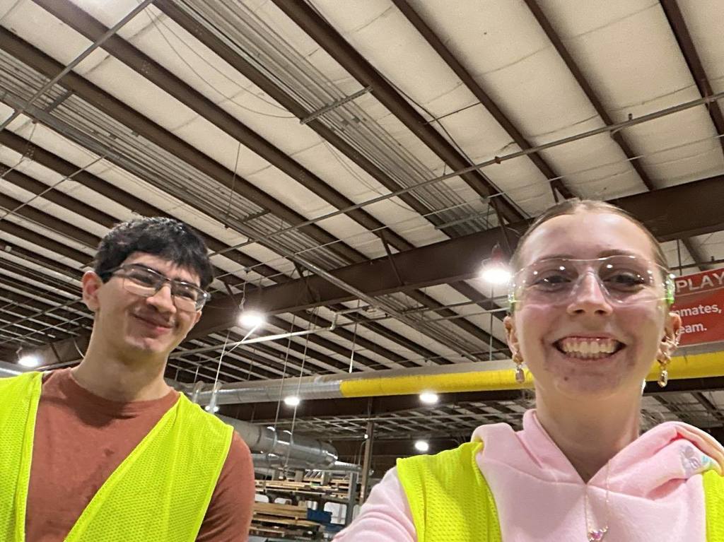 Two high school students, a boy and a girl, wearing yellow safety vests and clear safety glasses, smile for a photo inside the Bishop Fixtures and Millwork manufacturing facility.