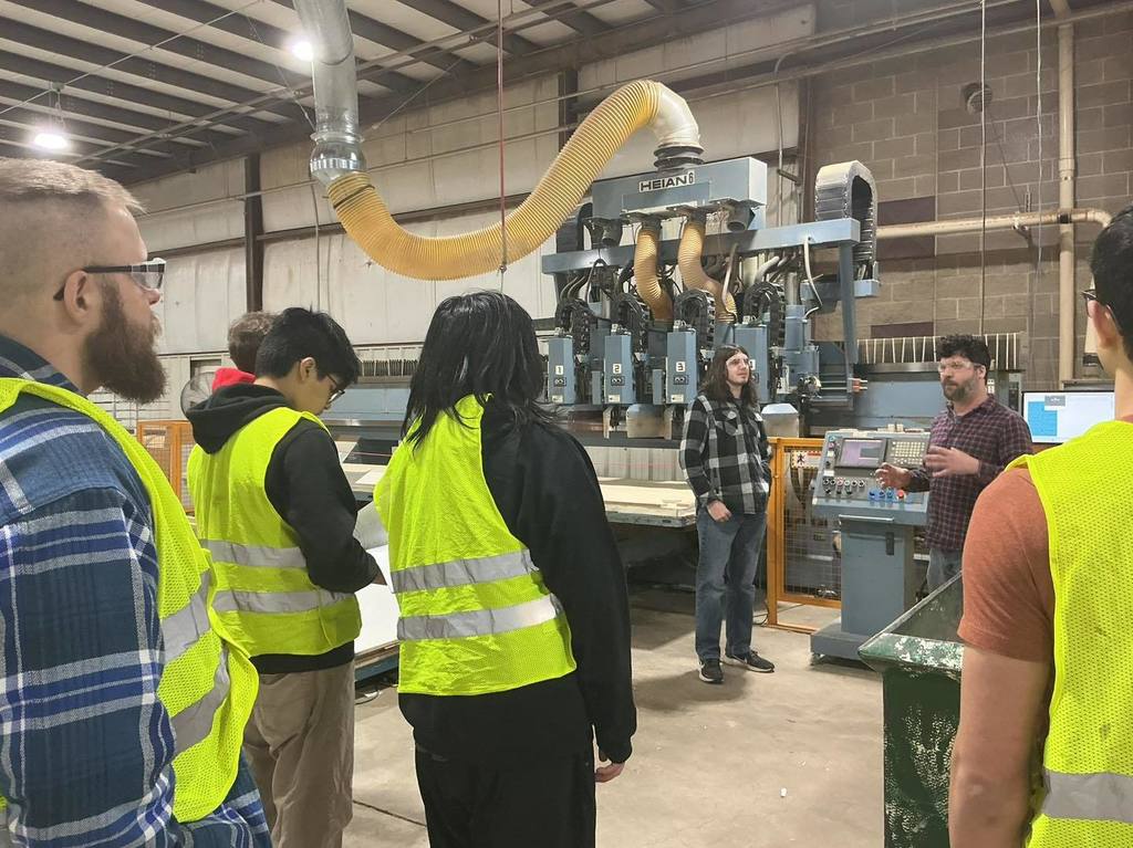 A group of high school students in yellow safety vests and a teacher in a blue plaid shirt stand in a workshop at Bishop Fixtures. They are listening to a staff member explain the operation of a large industrial CNC machine with a prominent yellow vacuum hose.