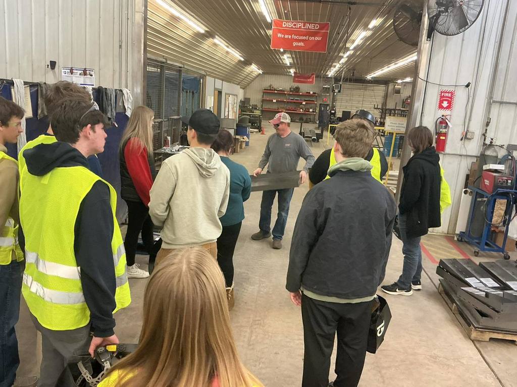 A group of Unity High School students in yellow safety vests stand in a circle in the Bishop Fixtures workshop. They are listening to an instructor in a gray shirt who is holding a long, flat metal component from a toolbox kit.