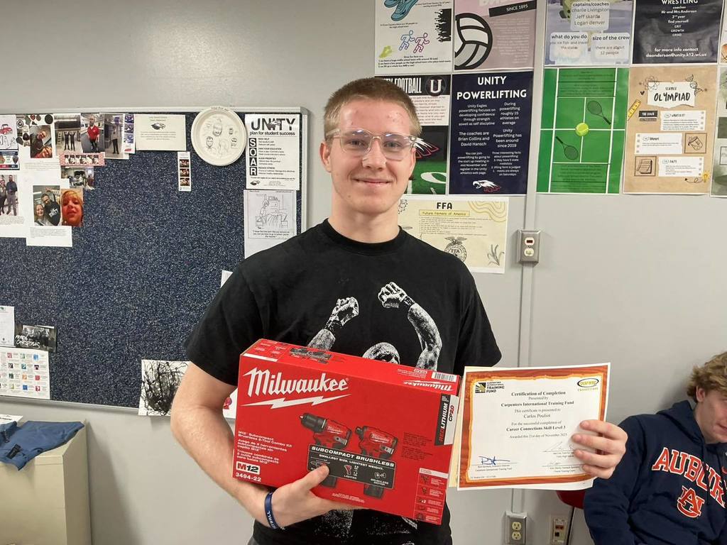 A Unity High School Residential Construction student wearing a black t-shirt and safety glasses smiles while holding a red Milwaukee power tool kit and his "Certification of Completion" certificate. He is standing in a classroom decorated with school posters.
