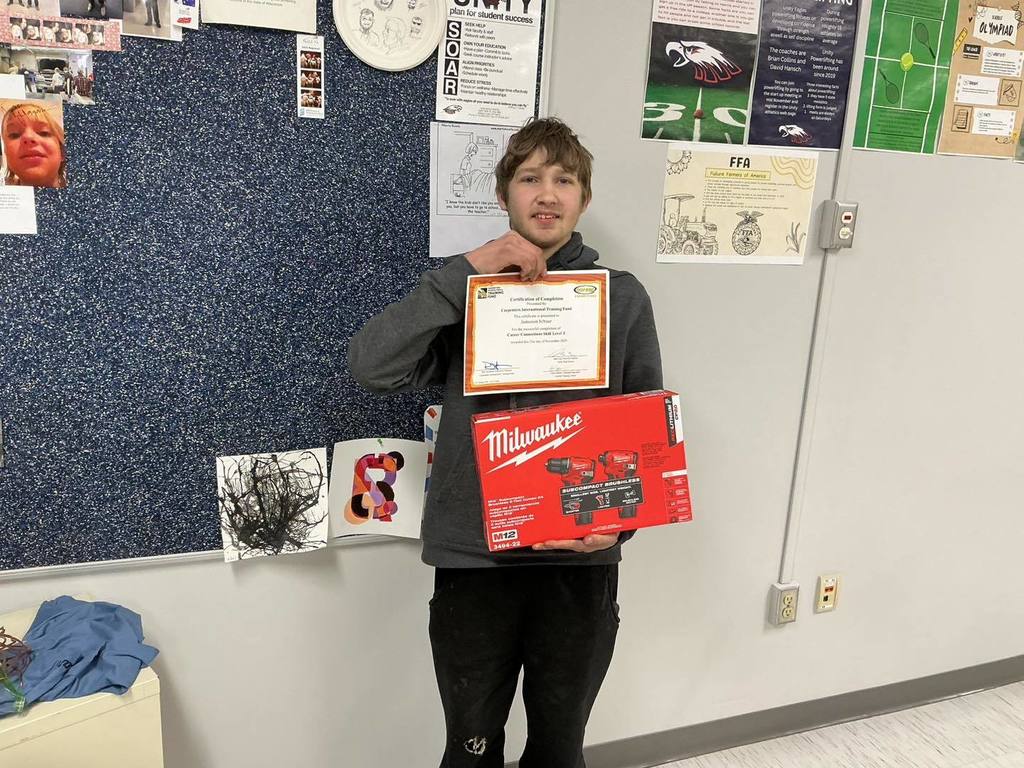 A Unity High School Residential Construction student smiles while holding a "Certification of Completion" from the Carpenter’s International Training Fund and a red Milwaukee power tool kit. He is standing in a classroom with student success posters and project photos on the wall behind him.