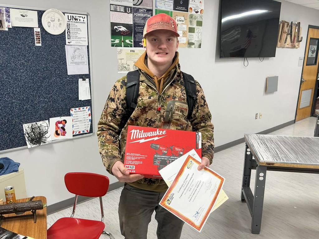 A Unity High School Residential Construction student wearing a camouflage jacket and an orange baseball cap. He is smiling while holding a red Milwaukee power tool kit and several "Certification of Completion" documents in a classroom setting.