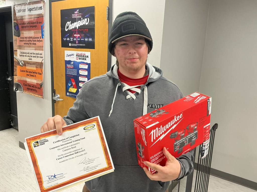 A Unity High School Residential Construction student wearing a black beanie and gray hoodie holds a "Certification of Completion" from the Carpenters International Training Fund and a red Milwaukee power tool box in a classroom.
