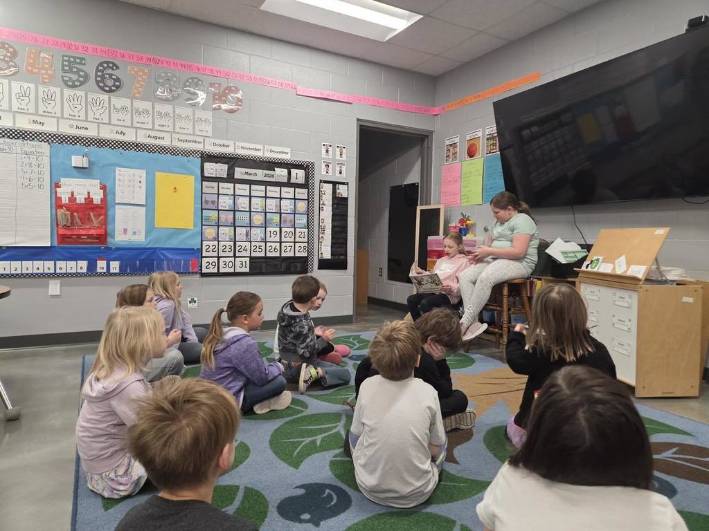 Two young students sitting at the front of a classroom reading a book aloud to a group of classmates sitting on a colorful rug.