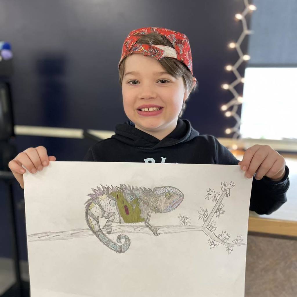 A smiling third-grade student named Archer holding up his detailed colored pencil drawing of a chameleon on a branch.